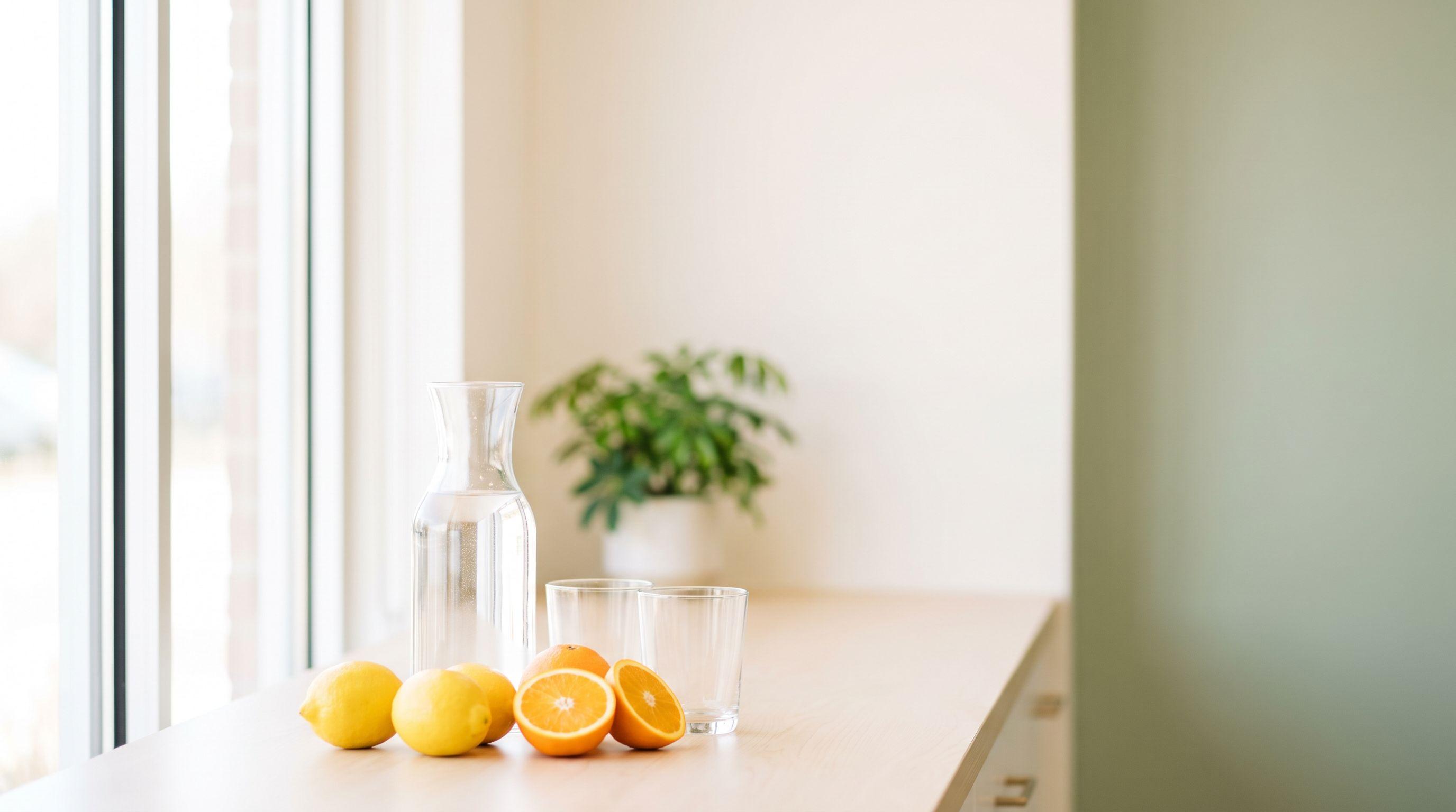 A glass carafe of water, two empty glasses, lemons and halved oranges on a pale wood clinic counter in soft daylight, with a green plant behind.