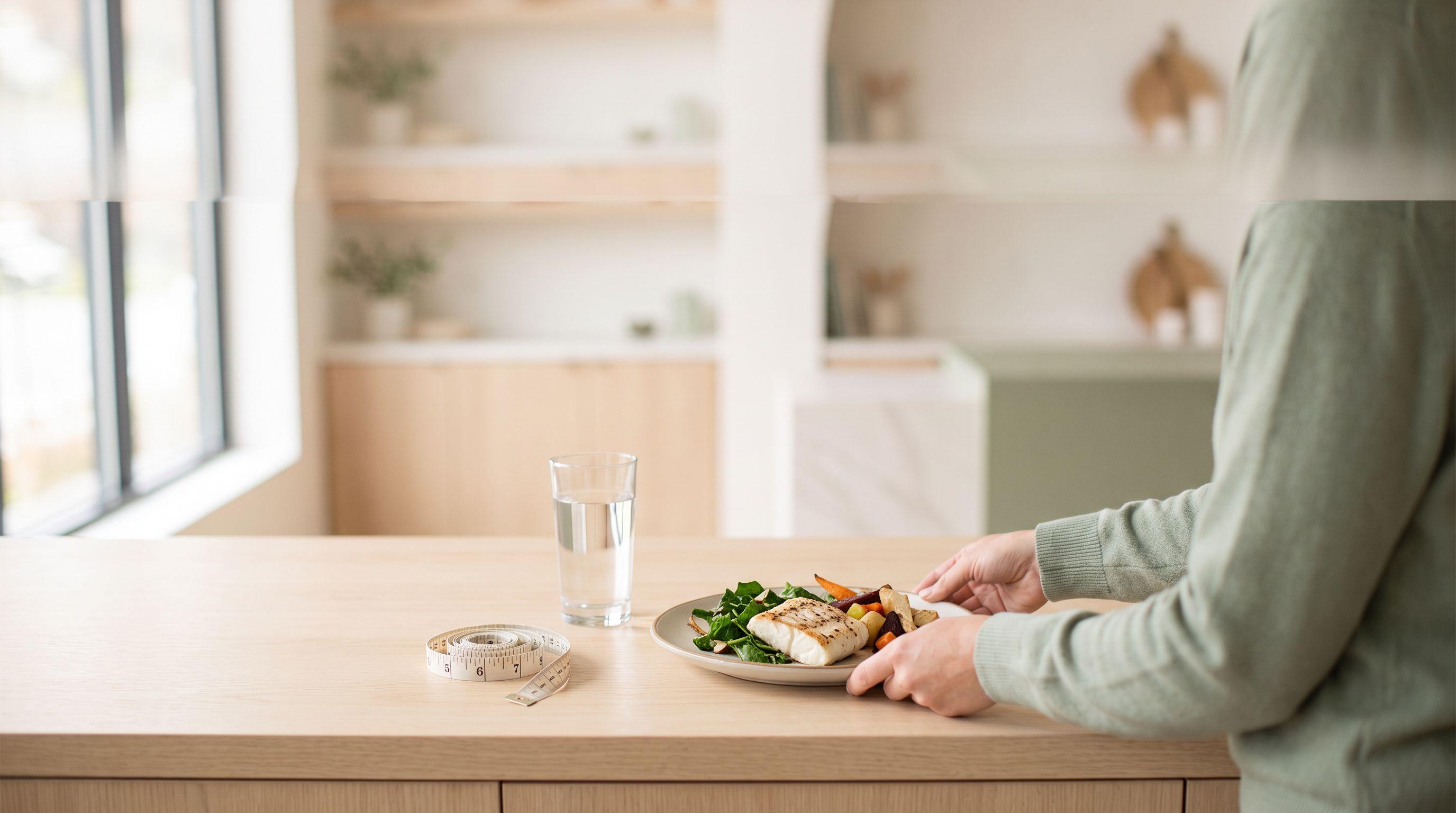 A simple plate of grilled fish, greens and roasted vegetables beside a glass of water on a pale wood kitchen counter, with a cloth tape measure coiled nearby in soft daylight.